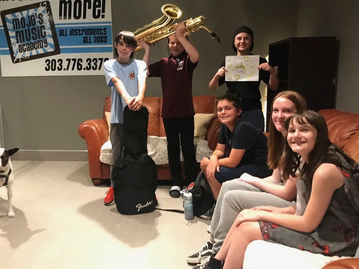 Six smiling Mojo's Music Academy students in the lobby with a baritone saxophone, a guitar and a hand-drawn picture