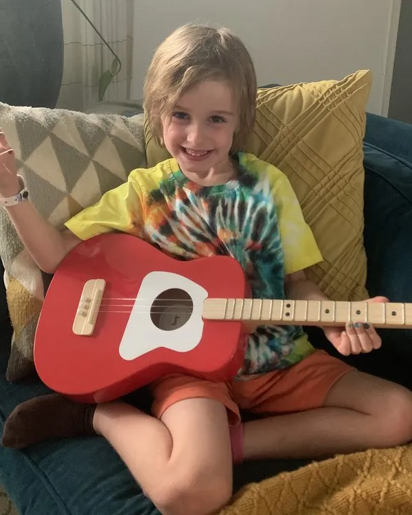A smiling young child playing a red 3-string guitar sized for small hands
