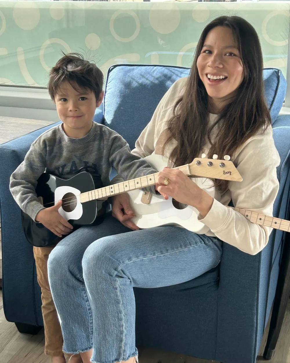 A mom and her young son smiling together, each holding a 3-string guitar sized for small hands